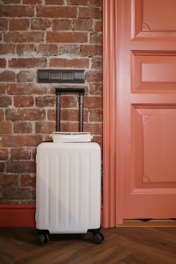 hero-img-01 Compact white suitcase against a rustic brick wall beside a coral door, perfect for travel themes.